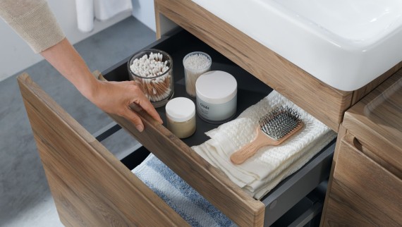 O mână deschide unul dintre sertarele dulapului de lavoar Geberit în design din lemn natural O mână deschide unul dintre sertarele dulapului de lavoar Geberit în design din lemn natural