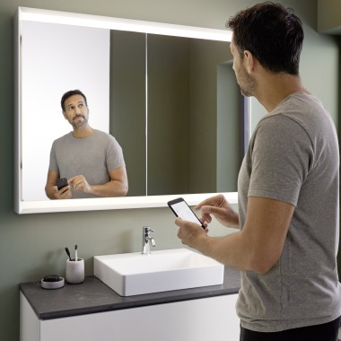Un bărbat controlează iluminarea dulapului cu oglinzi Geberit ONE cu ajutorul smartphone-ului său (© Geberit)